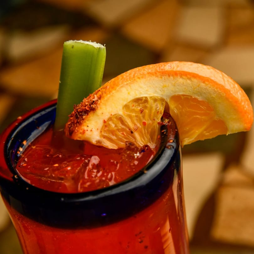 A vibrant red Michelada cocktail in a blue-rimmed glass, garnished with a celery stick and orange slice.