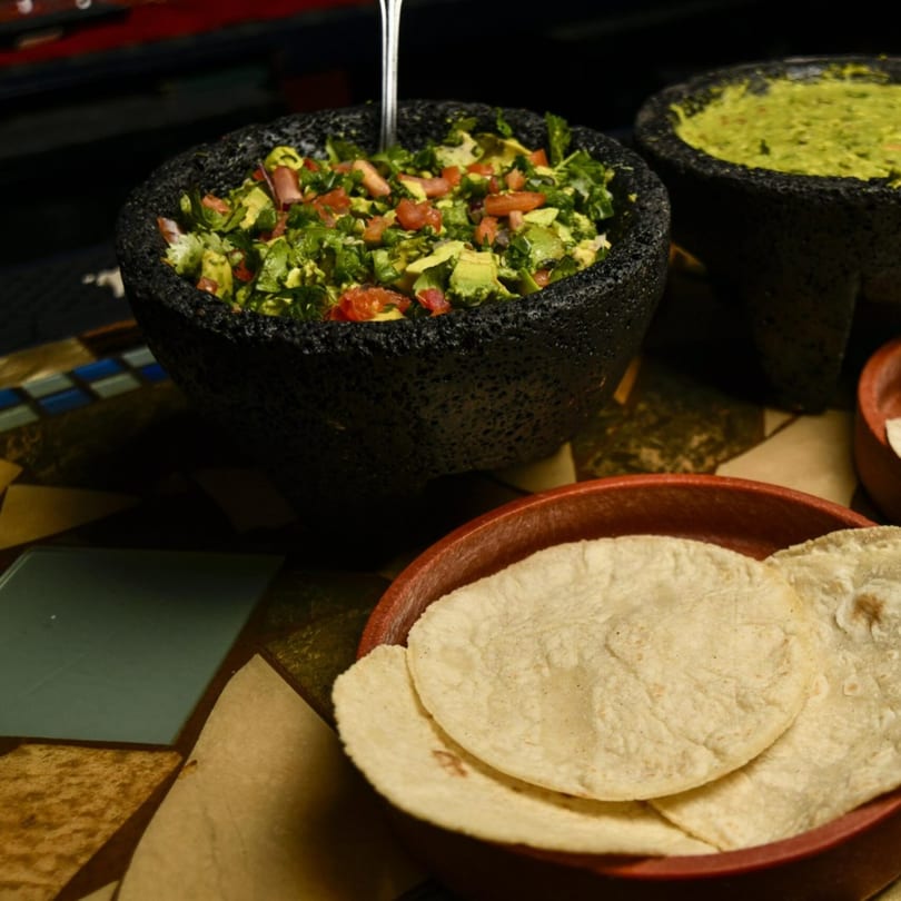 Fresh guacamole with tomatoes and cilantro in a molcajete, with tortillas in a red bowl.