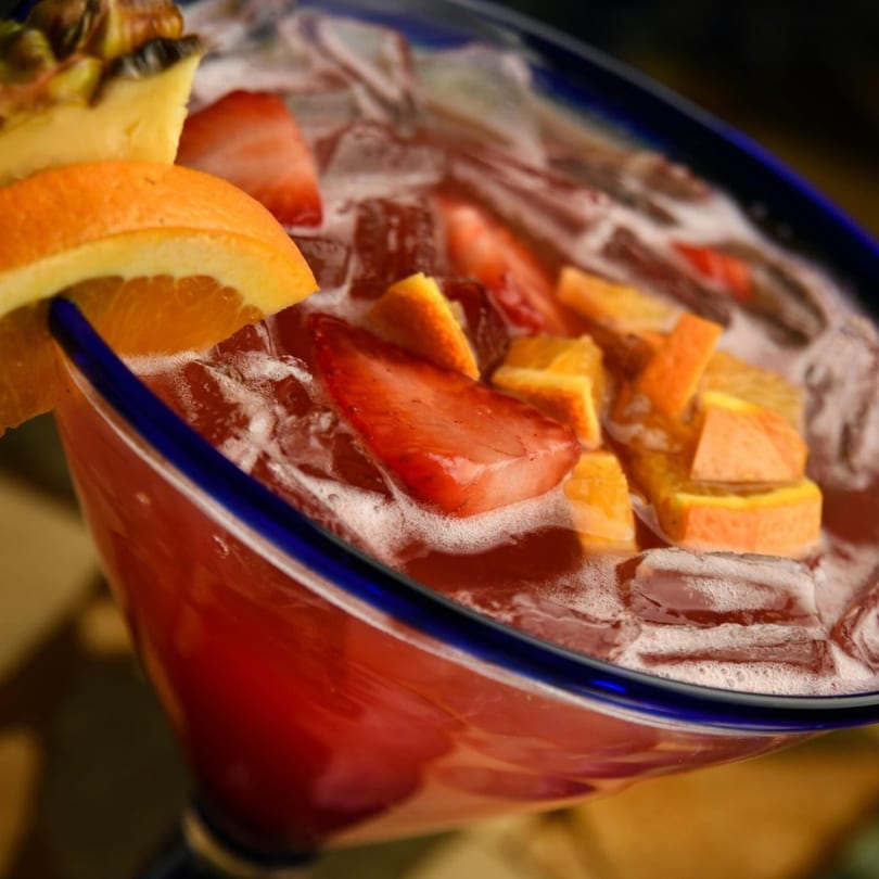 A vibrant red margarita with ice, garnished with orange, pineapple, and strawberries in a blue-rimmed glass.